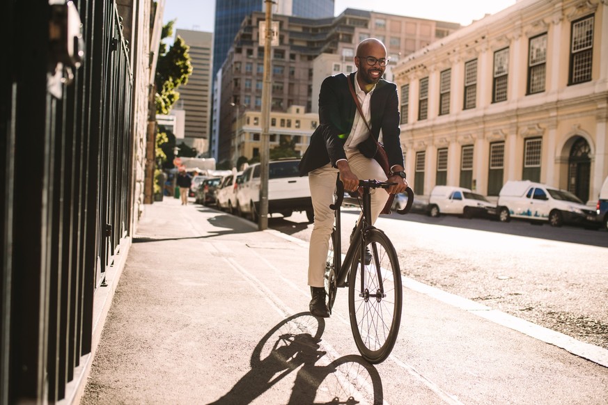 Young smiling businessman going to work by bike. Man in suit riding bicycle on sidewalk in morning.
