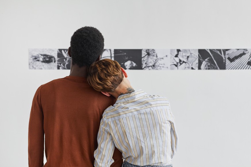 Back view portrait of mixed-race couple embracing while looking at paintings at modern art gallery exhibition, copy space