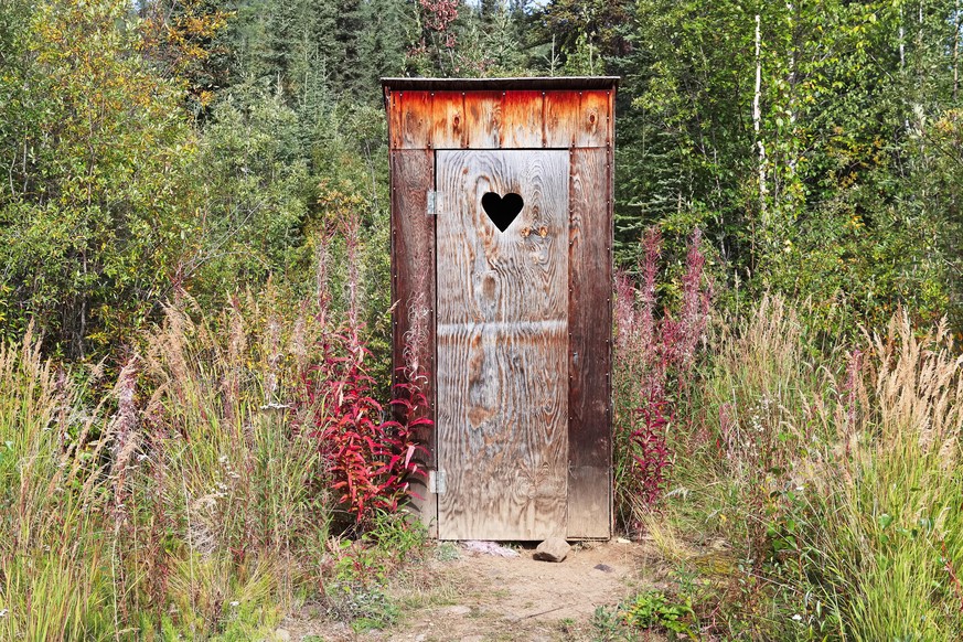 An outhouse in a wooded area with a heart window.