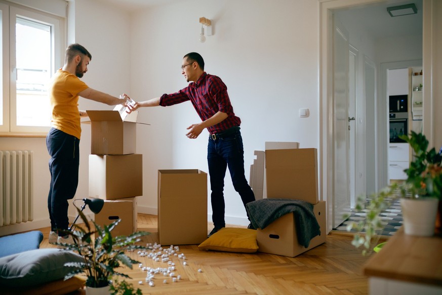 Gay couple in the empty apartment opening cardboard boxes and taking vase out together