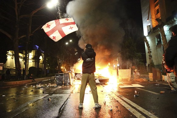 09.03.2023, Georgien, Tiflis: Ein Mann schwenkt eine georgische Nationalflagge vor einer brennenden Barrikade in der Nähe des georgischen Parlamentsgebäudes. Die Polizei in der georgischen Hauptstadt  ...
