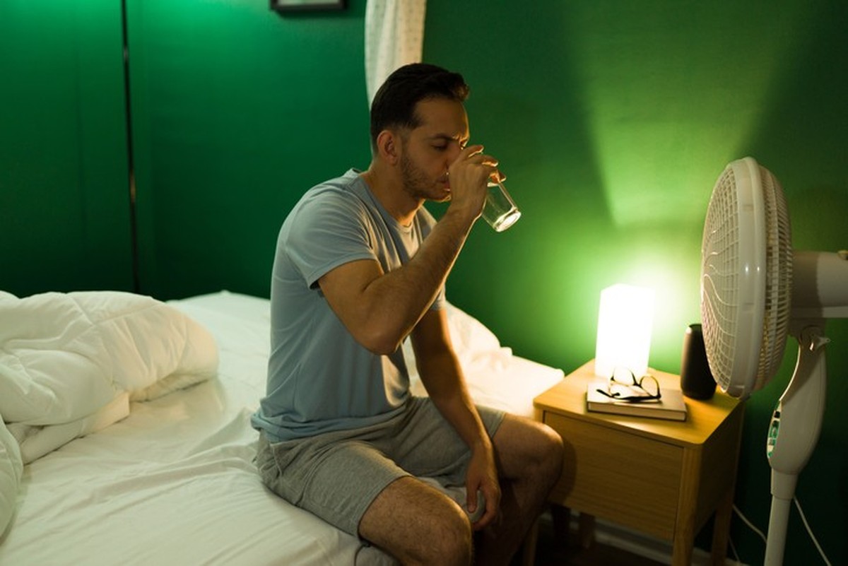 Hot night. Thirsty young man drinking water in front of a fan while sitting up in bed