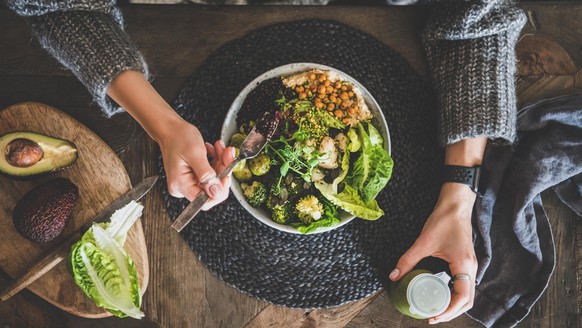 Healthy dinner, lunch setting. Flat-lay of vegan championship game or Buddha bowl with hummus, vegetable, salad, beans, couscous and avocado, green smoothie and woman's hands over wooden table, top vi ...