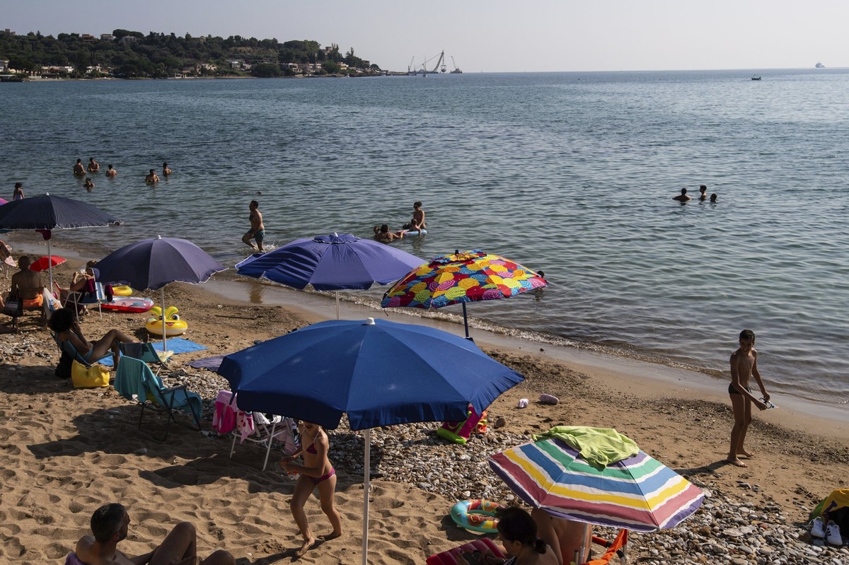 22.06.2025, Italien, Porticello: Touristen und Einheimische sonnen sich am Strand, während im Hintergrund der Rumpf der Superjacht «Bayesian» geborgen wird. Die als unsinkbar deklarierte Megajacht war ...