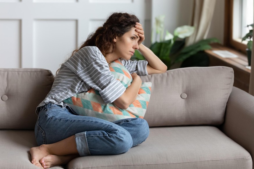 Frustrated young lady sitting on sofa, cuddling pillow, looking away at window. Lost in thoughts unhappy stressed millennial woman regretting of wrong decision, spending time alone in living room.