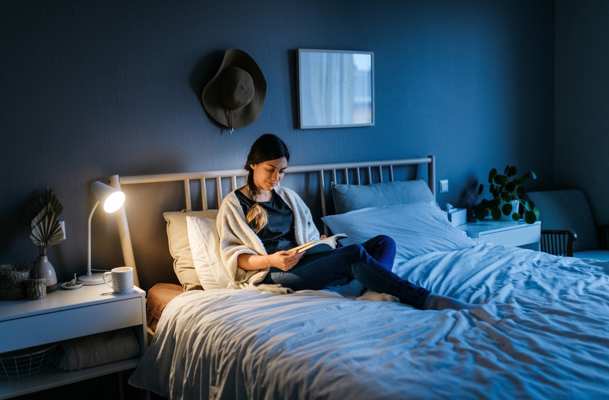 Young woman lying on the bed and reading a book at night. The room is dark and blue and the light on the night stand is illuminating the book. Bedroom, horizontal photo