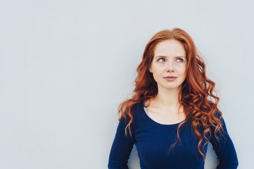 Attractive young woman standing pondering a problem looking up with a contemplative expression against a white exterior wall with copy space