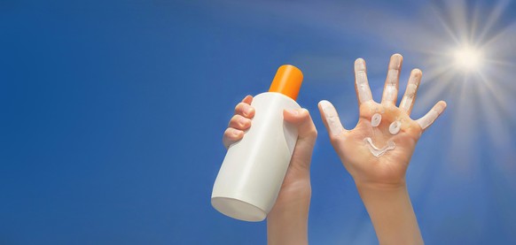 Positive symbol drawing by sunscreen on open hand. Another hand holding a bottle of sun cream. Blue sky, beams of sun background. Protection of sun. Copy space.