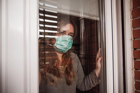 A young woman in quarantine wearing a mask and looking through the window