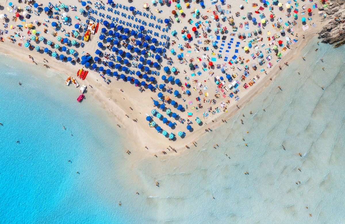 Aerial view of beautiful beach with white sand, colorful umbrellas, swimming people in blue sea at summer sunny day. La Pelosa beach, Sardinia, Italy. Top drone view of sandy beach, transparent water  ...