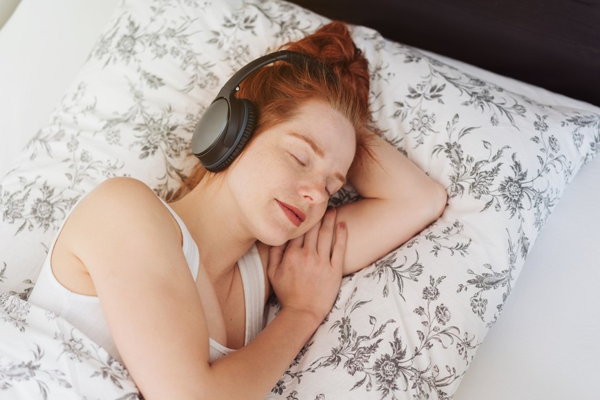 Young woman in white shirt sleeping on her side with black overhead headphones on head, listening to music or relaxing audio. Viewed in close-up from above