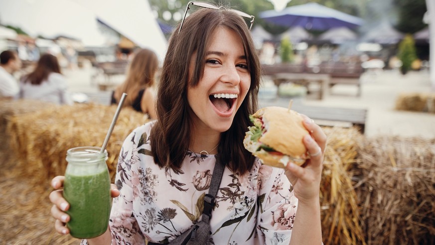 Stylish hipster girl in sunglasses holding delicious vegan burger and smoothie in glass jar in hands at street food festival. Happy boho woman holding burger and drink in summer street