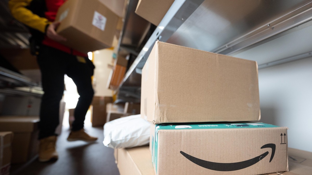 Ein Paket des Online-Händlers Amazon steht in einem DHL-Transporter für die Paketzustellung. Im Hintergrund sortiert ein DHL-Mitarbeiter seine Pakete für die Zustellung.