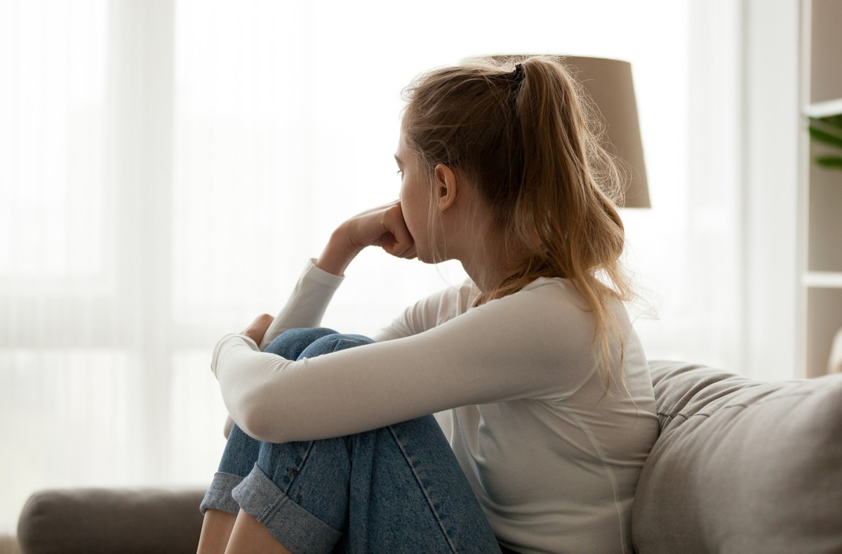 Side view young woman looking away at window sitting on couch at home. Frustrated confused female feels unhappy problem in personal life quarrel break up with boyfriend or unexpected pregnancy concept