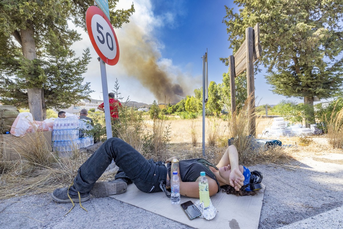 25.07.2023, Griechenland, Gennadi: Eine Helferin, die seit Tagen im Einsatz ist, hat sich zum Ausruhen am Stra�enrand auf ein St�ck Pappe gelegt, im Hintergrund die Rauchfahne eines neu ausgebrochenen ...