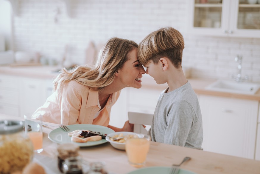 Emotionale junge Frau verbringt Zeit mit ihrem Sohn in der Küche und lächelt, während sie sein Gesicht mit ihrer Nase berührt