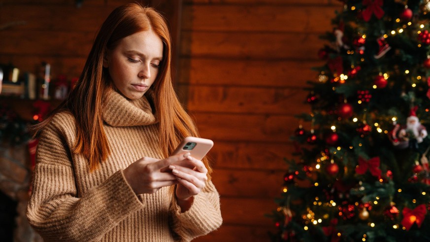 Sad young woman typing message on mobile phone while self-isolating during the winter holidays at room with festive interior. Concept of celebrating Christmas and New year at alone while lockdown.
