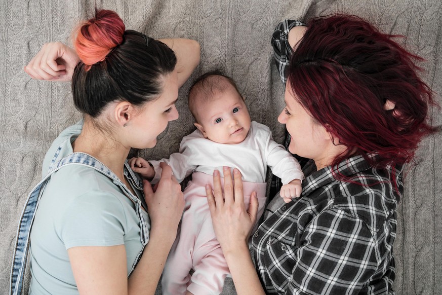 Two young women in casual clothes and with pink hair, a lesbian homosexual couple, lying on a rug with a child. Same-sex marriage, adoption.