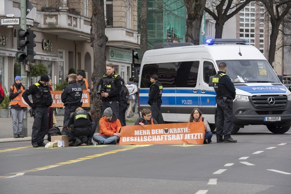 05.04.2023, Berlin: Aktivisten der Gruppe &quot;Letzte Generation&quot; haben sich auf der Neuen Kantstraße auf einer Kreuzung festgeklebt. Die Polizei sichert den Bereich, während eine Polizistin beg ...