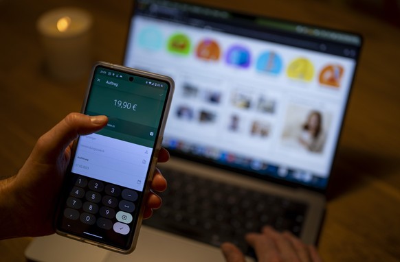 ARCHIV - 27.02.2023, Berlin: Ein Mann �berweist in einer Banking-App auf einem Smartphone Geld, w�hrend im Hintergrund auf einem Laptop die Seite eines Online-Handels angezeigt wird. Ob Hotelbuchung,  ...