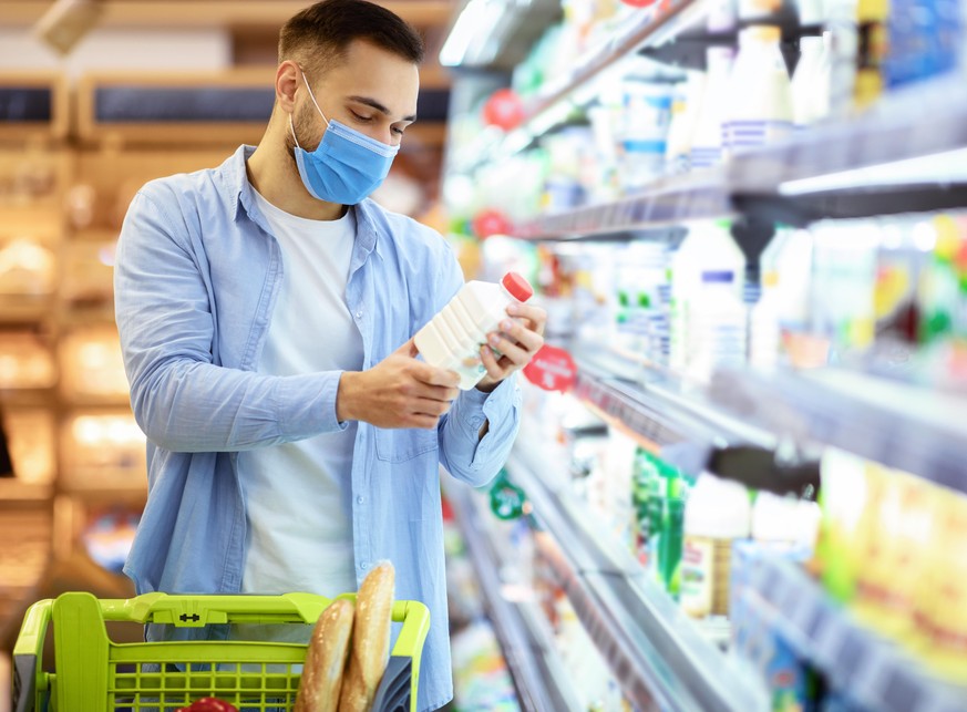 Choosing Milk. Young man in disposable medical surgical mask holding bottle of milk or yoghurt from the fridge, looking at dairy products, standing near refrigerator aisle, checking expiry date