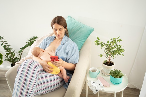 Young beautiful mother sitting in comfortable chair with feet on a footrest, breastfeeding her newborn baby, looking at smartphone. Smiling mom having peaceful bonding time with her baby. || Modellfre ...