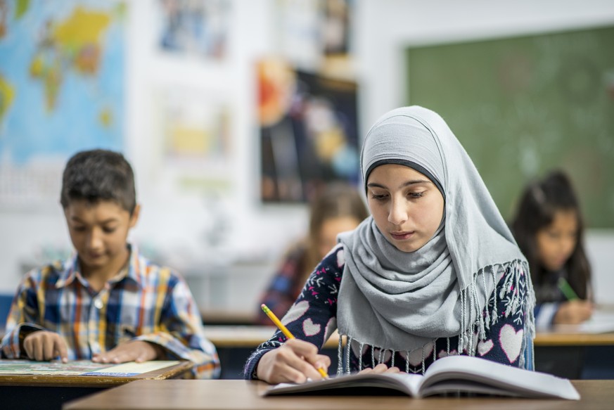 A multi-ethnic group of elementary school students are indoors in a classroom. They are wearing casual clothing. A Muslim girl wearing a head scarf is writing in her notebook.