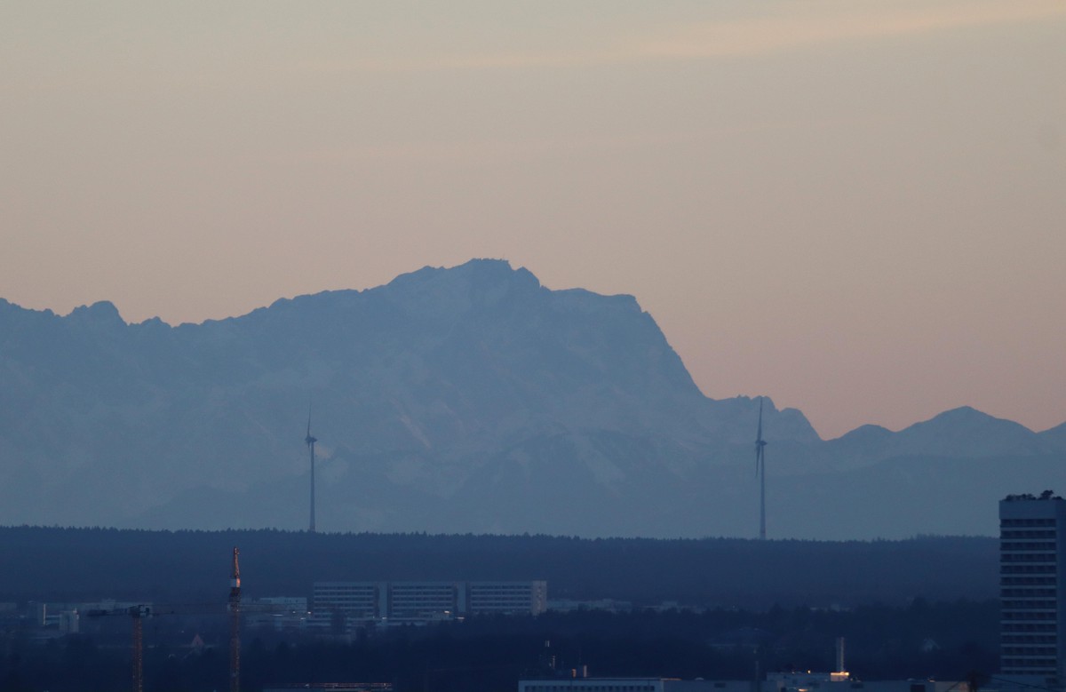 München, Bayern, Deutschland 12. Februar 2022: Ein Wintertag in der Landeshauptstadt München. Hier der Blick vom Olympiaberg auf die Stadt, zwei Windrädern und dem Massiv der Zugspitze