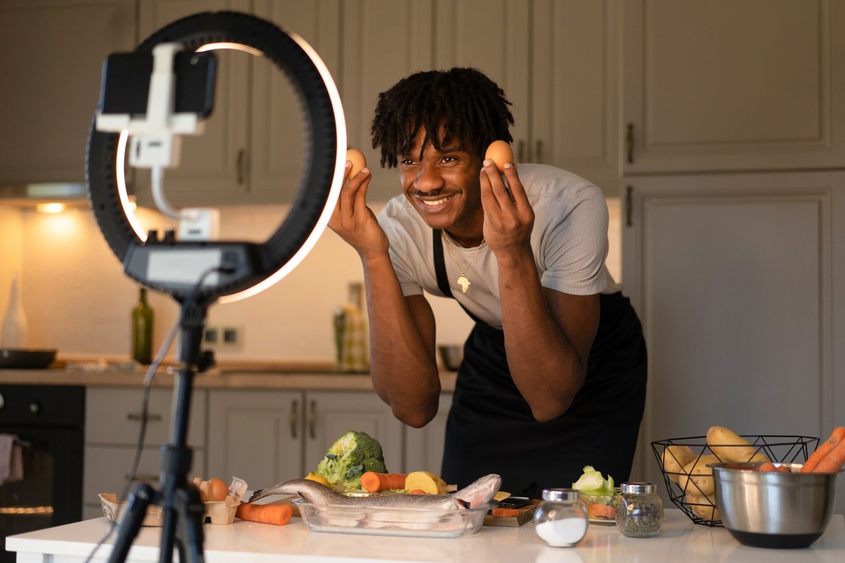Using a selfie LED ring light to hold his mobile while recording a video, this male cook is demonstrating the recipe for a cooking video using the ingredients spread out on the table before him