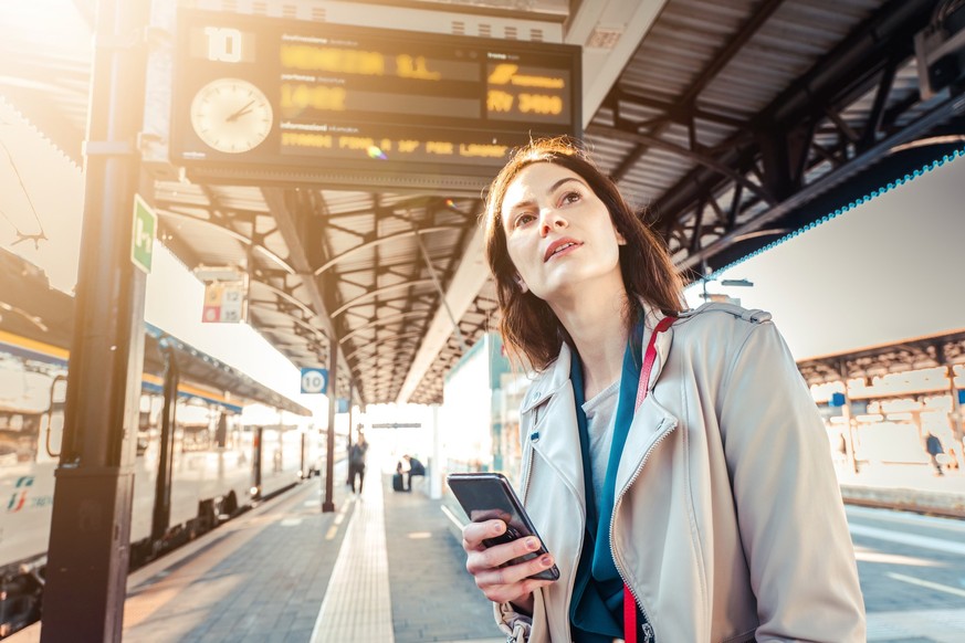 Young woman with departure times behind her waiting for her train while holding her mobile phone - Woman looking at the clock in the train station while her train is delayed - Transportation and urban ...