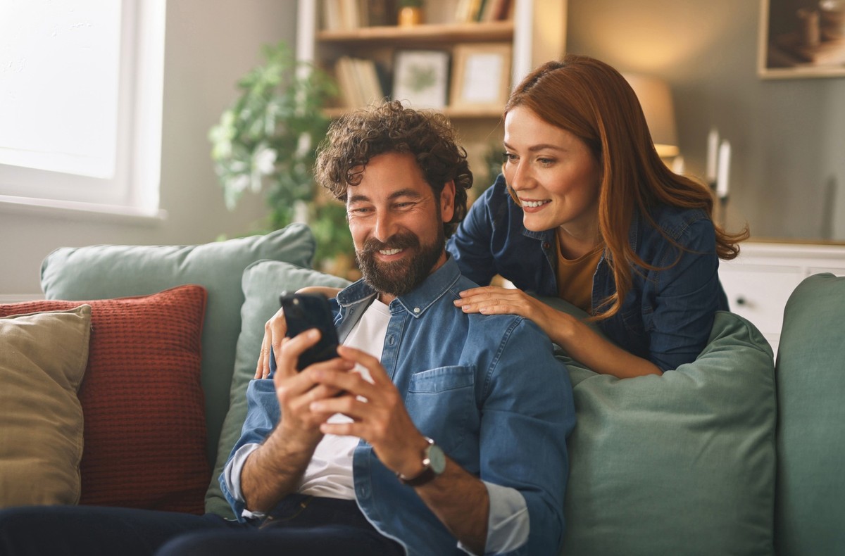 Couple shares a joyful moment in their new living room, smiling as they browse their smartphones, surrounded by comfortable decor and soft lighting.