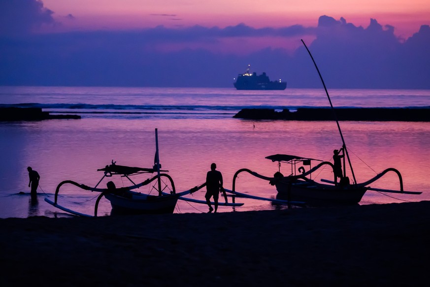 ARCHIV - 14.11.2022, Indonesien, Nusa Dua: Männer befestigen vor Sonnenaufgang am Tag vor Beginn des G20-Gipfels am Strand von Nusa Dua Boote, wobei im Hintergrund ein Schiff der indonesischen Marine  ...
