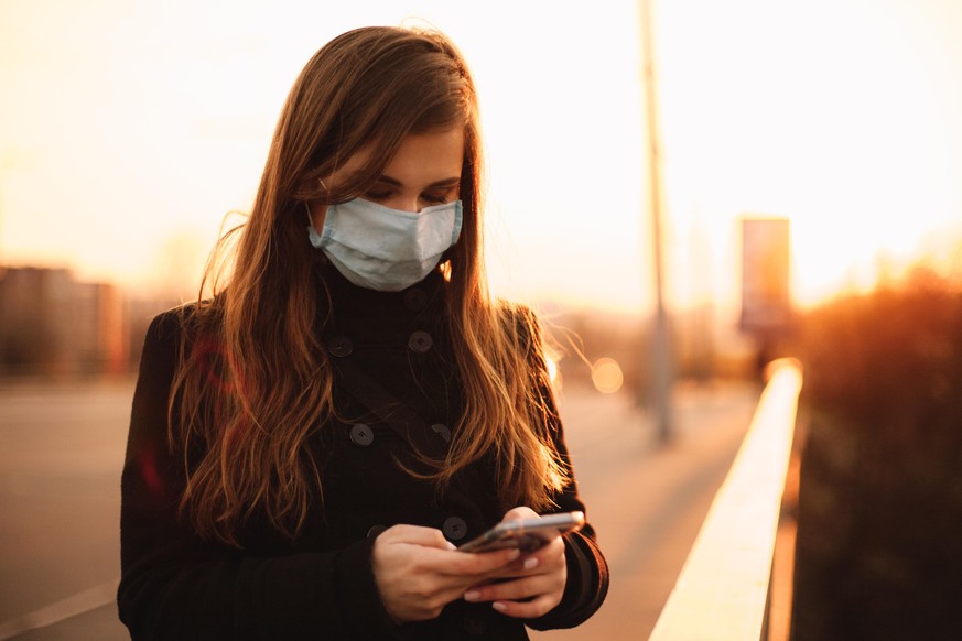 Young woman wearing protective face medical mask using smart phone while standing on bridge in city at sunset