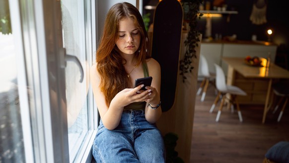 Caucasian teenage girl, sitting near a window and using mobile phone