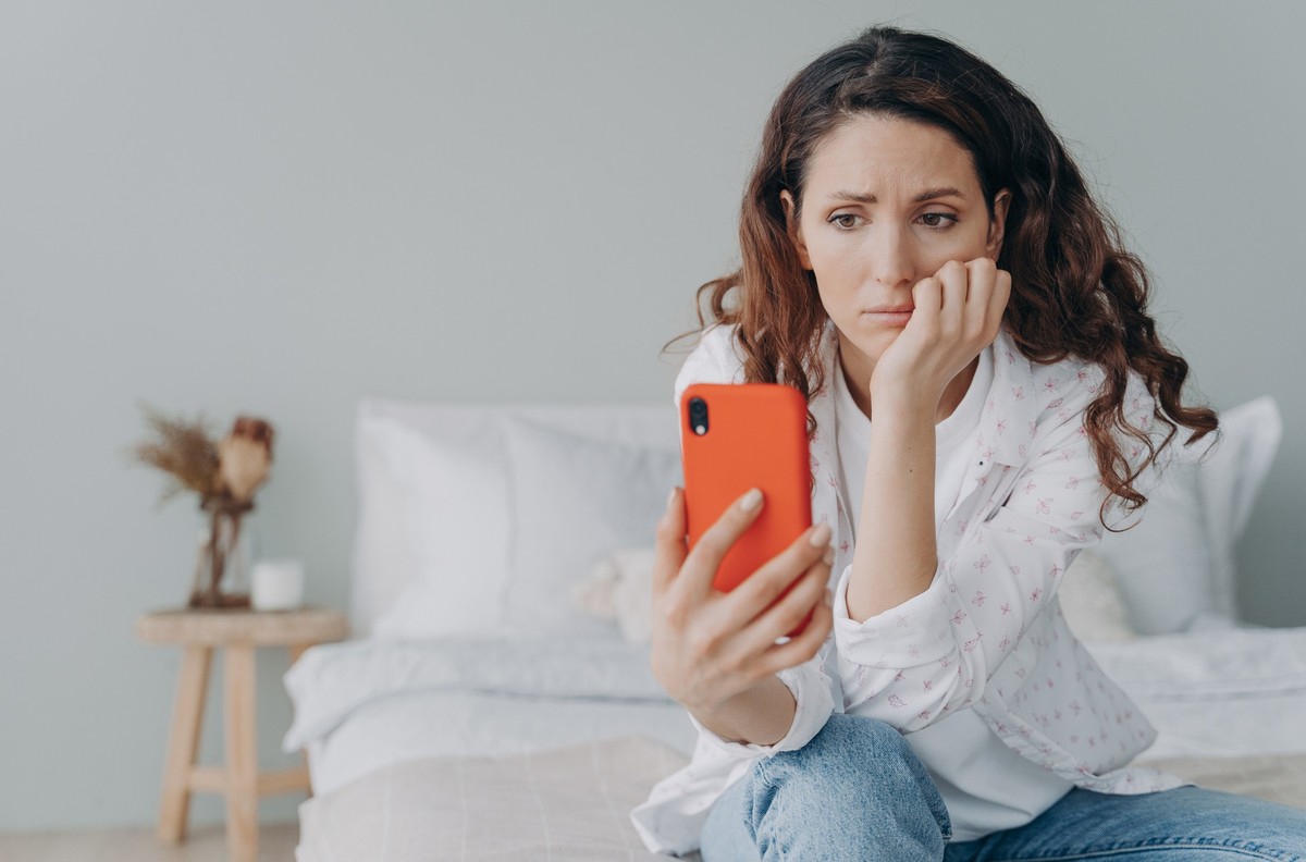 Stressed caucasian girl is reading morning news looking at smartphone display. Young woman is chatting on phone or browsing internet at home in her bedroom. Lady is doom scrolling.