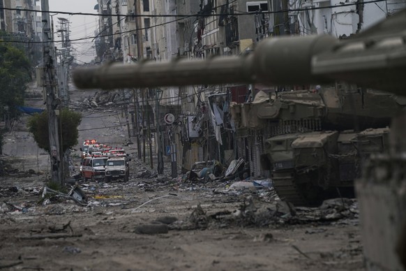 22.11.2023, Palästinensische Gebiete, Gaza-Stadt: Ein Konvoi mit Krankenwagen fährt durch eine von Trümmern übersäte Straße in der Nähe eines Panzers der israelischen Streitkräfte während einer Bodeno ...