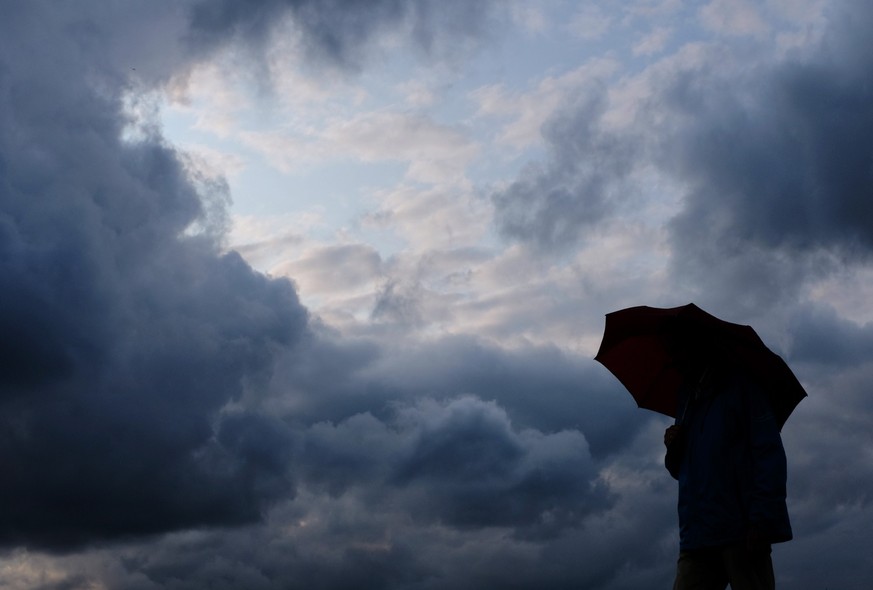 ARCHIV - 12.07.2019, Nordrhein-Westfalen, Duisburg: Ein Mann geht mit einem Regenschirm vor aufziehenden dunklen Wolken spazieren. Nach ersten sonnigen Frühlingstagen schwenkt das Wetter in Nordrhein- ...