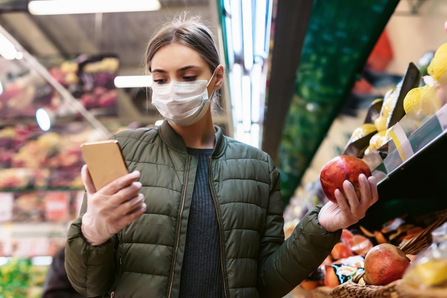 A young woman in a disposable face mask is checking a shopping list on a smartphone in a supermarket. Social distancing during the pandemic of the coronavirus Covid-19