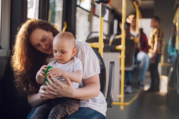 Young plus size mother in casual wear holding little toddler boy in her lap while riding a bus together in sunny day. Mom and her child enjoying a bus journey together.