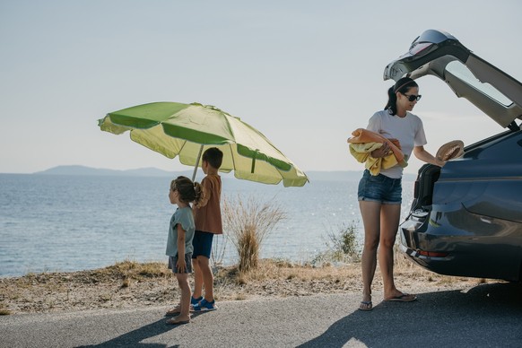 Mother with her son and daughter arrived at the sea, mother is taking towels from a car trunk for the beach.