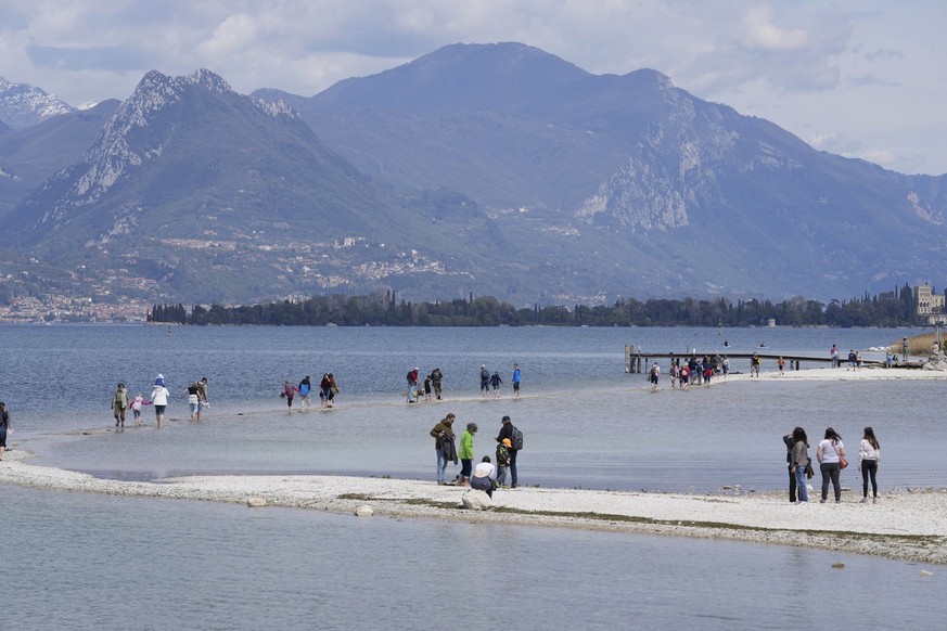 14.04.2023, Italien, Manerba del Garda: Touristen gehen über einen Streifen Land zwischen dem Festland und der Insel San Biagio, am Gardasee. Die anhaltende Trockenheit und Wasserknappheit machen Ital ...