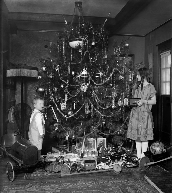 Abundant toys are underneath a traditionally decorated Christmas tree, c. 1920. A boy and girl stand beside holiday tree in their home. Modern motor vehicles are among the presents under the tree BSLO ...