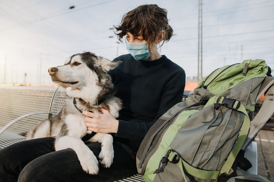 Casual Woman with backpack is waiting for the train with her dog on the platform and is wearing a face mask because of the coronavirus. Lovely und happy friendship with your pet.
