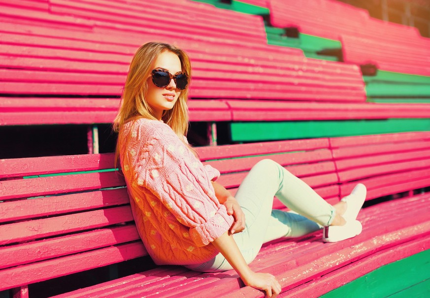 Portrait of beautiful young blonde woman sitting on bench wearing sunglasses, sweater in the city park