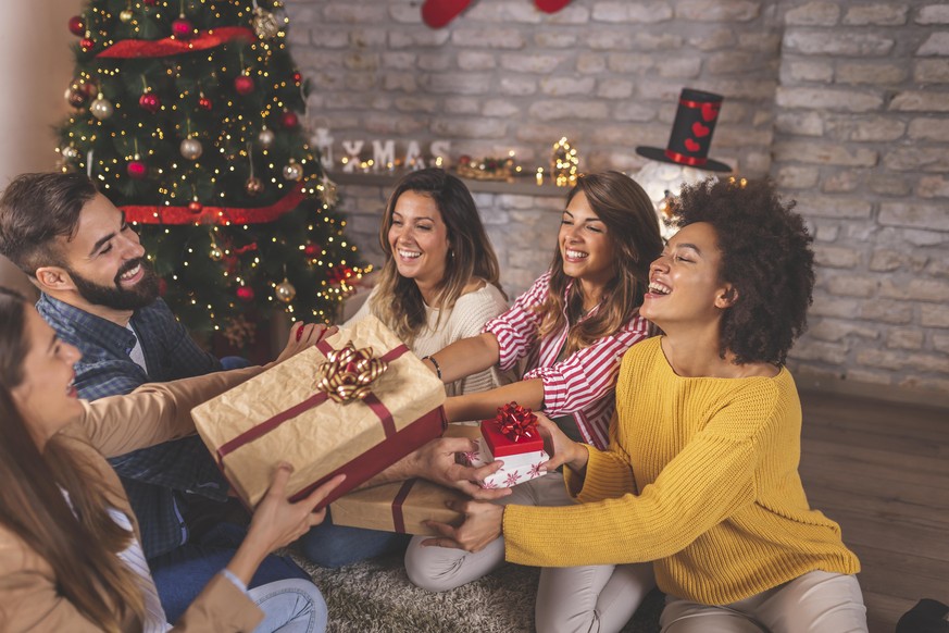 Group of friends sitting by nicely decorated Christmas tree, celebrating Christmas at home, exchanging presents and having fun