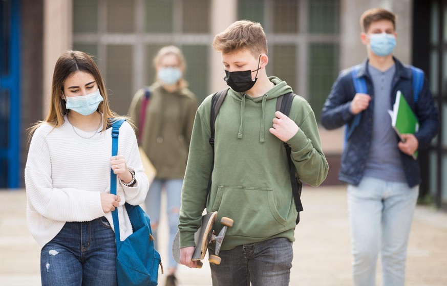 Teen girl in protective face mask walking with her classmate outside college building on sunny day