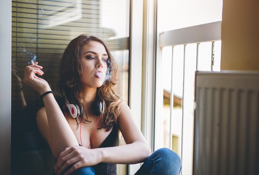 Woman with headphones sitting next to the window and smoking a cigarette.