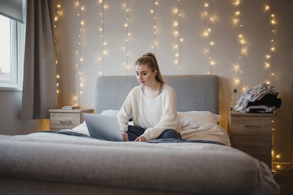 Young woman is sitting on her bed using a laptop.