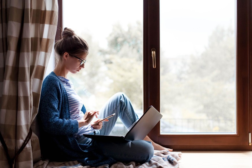 Working from home. Young woman using laptop, phone, Internet. Freelancer workplace in living room office on windowsill. Distance learning, isolation, female business, shopping online. Lifestyle moment