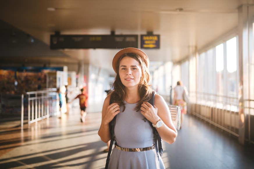 Theme tourism travel. Young beautiful Caucasian woman in dress and hat with backpack tourist mat in terminal station in a long walkway, the schedule board, the gate of the departure and the plane.